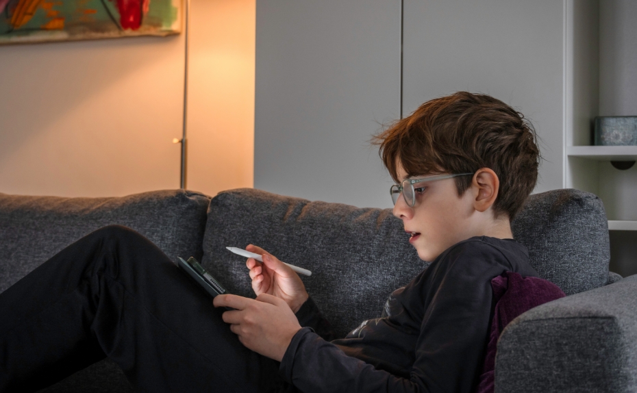 Junge mit Brille sitzt auf Sofa mit Tablet auf dem Schoss. 